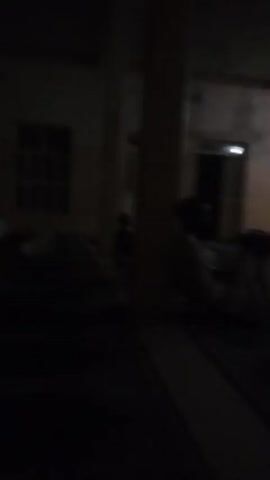 This video shows the interior of a dimly lit room, possibly a mosque or prayer hall, with green carpeted floors marked with rows of white lines. Several people are present, some are kneeling or sitting on the floor in prayer, while others are standing. The lighting is low, with some illuminated ceiling lights and a fan visible. The walls are a light color, and there are windows and doorways visible in the background. The overall atmosphere is quiet and contemplative.