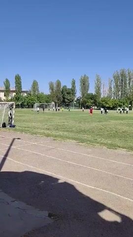 A group of young people are playing soccer on a grassy field. The field has a running track around it. The sky is clear and blue. There are trees and buildings in the background. The video is shaky, suggesting it was filmed by someone walking or running.