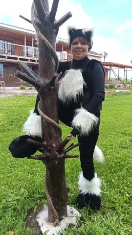 The video shows a woman dressed in a black cat costume with white furry accents on her ears, chest, wrists, feet, and tail. She is posing next to a wooden structure that resembles a tree with branches. The structure is wrapped with a thick rope.

The woman is standing on a grassy area, with a wooden building in the background. The building appears to be a house or a lodge with a balcony and windows. There are some trees and greenery around. The sky is visible and appears to be bright, suggesting it is daytime.

The woman is smiling and looking at the camera. Her posture suggests she is posing for the picture. The overall atmosphere seems cheerful and playful, given the costume and the outdoor setting. There is no visible text in the video. The location is identified as Medvedevka, Russia.
