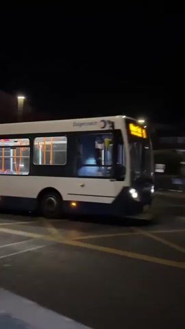 A white and blue bus with orange stripes is seen driving at night. The bus has illuminated windows and exterior lights. The bus appears to be a public transportation vehicle, possibly a city bus. The environment is dark, suggesting it is nighttime. The bus is on a paved road, and there are some faint yellow markings on the road surface visible in the foreground. The video appears to be captured from a stationary position, possibly from a sidewalk or an elevated viewpoint, as indicated by the perspective.