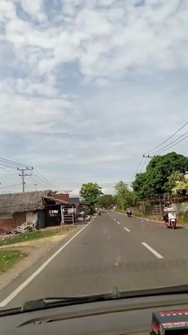 The video shows a view from inside a vehicle driving on a road. The road is paved and has white lines marking the lanes. There are trees and small buildings along the side of the road. Motorbikes and cars are visible driving on the road. The sky is cloudy.
