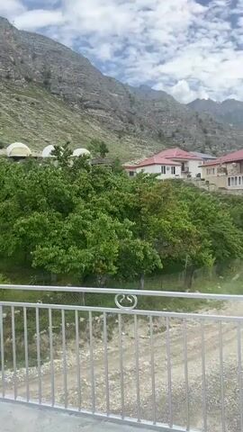 The video pans across a mountainous landscape with scattered buildings and vegetation. In the foreground, a white metal railing is visible. Beyond the railing, a dirt road winds its way up a hill. Several houses with red roofs are nestled amongst green trees on the hillside. Further in the distance, more houses and buildings can be seen, some appearing to be under construction. A large, imposing mountain range dominates the background, with some areas appearing barren and rocky, while others are covered in greenery. The sky is partly cloudy with a mix of blue sky and white clouds. There are no people visible in the video. The overall impression is of a rural village situated in a rugged, mountainous environment. The video then focuses on a body of water, possibly a river or lake, and a concrete structure that appears to be part of a dam or hydroelectric power station. Construction equipment like cranes are visible in the vicinity of the structure. The video ends with a close-up of the construction site.