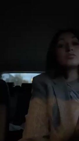 A young woman is seated in the back of a car. She wears a gray blazer and a gray t-shirt with a necklace. She has short, dark hair and is looking slightly upwards. The car interior is dark. A banana is held to her mouth, and she takes a bite from it. A blurry view of a street is visible through the back window.
