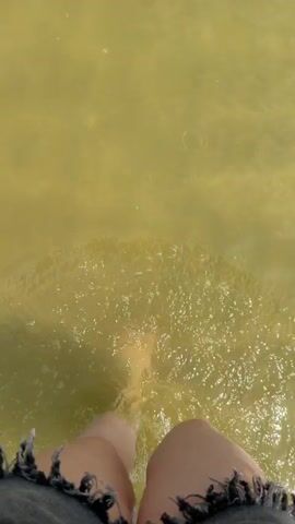 The video begins with a close-up of a person's bare foot splashing into murky, greenish water. The camera then pans up to show their leg, clad in denim shorts with frayed edges. The perspective shifts to a wider shot, showing the person, a male wearing a light-colored t-shirt and blue shorts, walking into the water. He is walking away from the camera, towards a vast body of water under a cloudy sky. In the distance, a jet ski is visible, creating a spray of water as it moves across the lake. The person continues to walk into the water, disappearing from view. The video ends with a view of the lake with the jet ski in the distance.