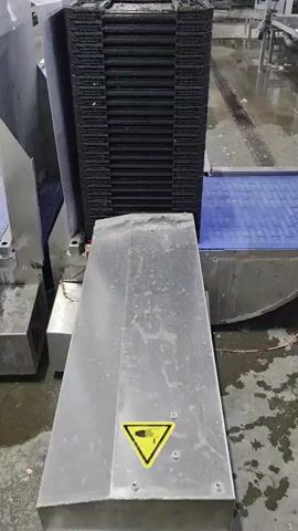 This is a top-down video of an industrial conveyor belt system in what appears to be a food processing facility. A stack of black plastic crates is being moved along a blue conveyor belt. The crates are stacked vertically and appear to be empty. The conveyor belt is moving from left to right in the frame. There is a metal chute or funnel directing the crates onto the conveyor belt, and a yellow caution sign depicting a hand with a pinch point warning is visible on the metal surface. The floor is wet and shows signs of regular cleaning. The overall environment is utilitarian and industrial.