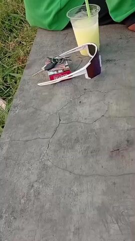 The video shows a close-up view of a concrete surface with a pack of cigarettes, a set of keys, and a pair of white sunglasses placed on top of the cigarettes. To the right of these items is a clear plastic cup filled with a pale yellow liquid, likely a beverage, with a green straw sticking out of it. In the background, out of focus, a person wearing a green shirt is partially visible. The environment appears to be outdoors, with some grass and foliage visible to the left. The concrete surface has a cracked texture.