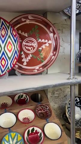 The video shows a close-up view of several decorative plates and bowls displayed on shelves in what appears to be a shop. The plates are round and feature intricate, colorful patterns, including geometric designs and floral motifs. Some plates have a dark background with bright, contrasting patterns, while others are reddish-brown with lighter colored designs. There are also smaller bowls, some of which have floral patterns painted inside. In the foreground of the second shelf, there are several small terracotta pots. The lighting suggests an indoor setting, likely a retail environment. There are no people visible in the video.