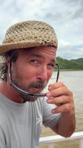A man wearing a woven straw hat and a light gray t-shirt is looking at the camera. He has a beard and is holding a pair of sunglasses in his right hand. He is winking at the camera. The background shows a body of water, possibly a river, with green hills in the distance under a cloudy sky. The man appears to be on a boat.