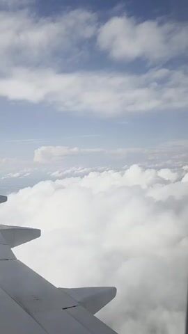 The video shows a view from an airplane window. The foreground features the wing of the airplane, extending from the lower left corner of the frame and angling upwards. The wing is white and appears to be made of metal. Beyond the wing, a vast expanse of white, fluffy clouds fills the mid-ground and extends into the background. These clouds are illuminated by sunlight, creating bright white masses. Above the clouds, the sky is a pale blue, with a few smaller, wispy white clouds scattered across it. The overall impression is of a serene, high-altitude flight over a sea of clouds.