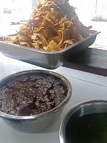 The video shows a variety of food items displayed in metal trays, likely in an outdoor or open-air setting. In the foreground, a large metal tray is filled with a mound of crispy, golden-brown fried snacks, possibly potato chips or plantain chips. Below this, another round metal container holds a dark, wet substance with white specks, possibly a sauce or a dish with seeds.

The video then pans to show other trays containing cooked food. One tray holds dark, rich-looking meat or stew with a liquid base. Another tray displays pieces of chicken or poultry coated in a green sauce, interspersed with some yellow elements. Further down, a tray is filled with chopped, dark-colored vegetables, appearing to be fried or stewed eggplant.

The final shots reveal two more trays. One contains a pile of what looks like small, dark, possibly fried food items, while the adjacent tray holds green, leafy vegetables or small pieces of food. In the lower section of the frame, a tray with sliced green vegetables, possibly cucumbers, and another containing small, brown items, possibly nuts or seeds, are visible.

The background suggests an outdoor environment with trees and a road. A white van is partially visible in the distance on the left, and a truck is seen on the right. The overall impression is of a food stall or a catering setup displaying a diverse selection of prepared dishes.