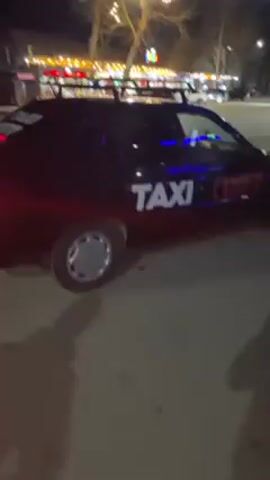 The video shows a man at night, likely on a city street, possibly near some shops or restaurants, based on the lights in the background. He is wearing a black jacket, a black hat, and looks directly at the camera. The camera is unsteady. There are cars parked on the street, and a car with its brake lights on pulls up behind him. The overall scene is dark, but the man is visible due to the available lighting.