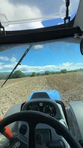 This is a first-person perspective video from inside a tractor cabin. The view is through the front windshield. The tractor is moving across a tilled field under a bright blue sky with scattered white clouds. In the distance, there are trees and rolling hills. The interior of the cabin shows the steering wheel, dashboard with gauges, and part of the tractor's blue hood. A windshield wiper is visible on the glass. The scene suggests agricultural work is in progress.