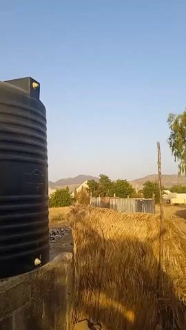 The video presents a static outdoor shot of a village-like setting, with a clear blue sky visible above. In the foreground, there is a tall, black water tank on the left side of the frame. Adjacent to the tank is a barrier constructed from straw or similar dry vegetation, creating a rustic fence-like structure. The ground is mostly bare, sandy soil. 

The background features a collection of buildings, some made of corrugated metal and others of a more traditional construction, possibly clay or mud. Trees are scattered throughout the scene, adding patches of green. In the distance, there are low, rolling hills or mountains under the clear sky. A silver car is parked along a road or path beyond the straw barrier. Overall, the scene gives the impression of a rural area, possibly in a hot or arid climate.