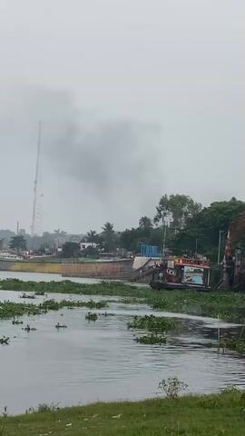 The video shows a river scene on a cloudy day. There are several boats docked along the riverbank, which is covered in vegetation. A tall communication tower is visible in the background, partially obscured by a large cloud of what appears to be smoke. The river is partially covered with aquatic plants.