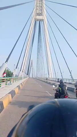 The video shows a person riding a motorcycle on a cable-stayed bridge. The camera is mounted on the front of the motorcycle, looking forward. The bridge has tall, white towers with many cables extending from them to support the roadway. There are other motorcycles on the bridge, and the sky is clear and blue. The bridge appears to be in a tropical or subtropical region, with green vegetation visible on the left side. There are safety barriers on the sides of the bridge.