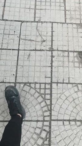 The video shows a first-person view of someone walking along a sidewalk. The sidewalk is made of gray paving stones. There is a road to the right, with cars parked along it. Buildings are visible on both sides of the road, including a white building with arched windows and a brown tile roof. The sky is overcast. The person walks along the sidewalk and then stops at a crosswalk.