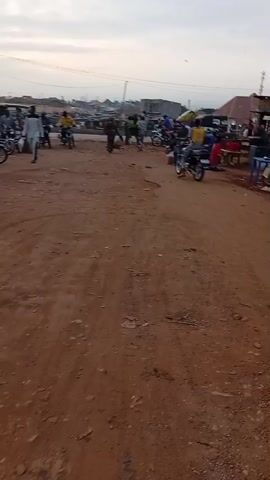 The video shows a wide-angle view of a busy dirt road area. Several people are visible, some standing and some sitting. Many motorcycles are seen passing by, some with riders and passengers. A few three-wheeled vehicles are also present. Along one side of the road, there are small stalls or shops with some individuals seated on chairs and benches nearby. The setting seems to be a marketplace or a local roadside area. The sky appears overcast and the light is somewhat subdued. The ground is mostly a reddish-brown dirt surface.