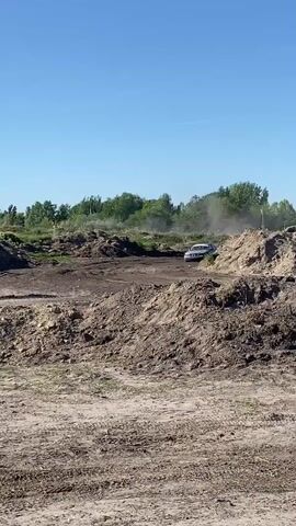 A silver sedan is driven aggressively over a dirt course, kicking up dust. The car crashes into a dirt mound, next to some flags, and then powers backwards away from the mound. The car has sponsor decals and racing numbers on its sides. The dirt course appears to be an open area with piles of dirt, grass, and trees in the background. The sky is blue and clear.