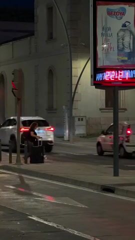 A person walks along a sidewalk with a suitcase at night. A digital billboard displays the temperature as 22 degrees Celsius. A white car drives past on the road. Another white car is parked further down the road. A traffic light is visible on the left side of the frame. The background consists of a building and streetlights.