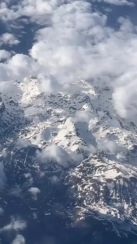 This video captures an aerial view of a vast, snow-covered mountain range, partially enveloped by white clouds. The perspective is from an aircraft, looking down onto rugged peaks and valleys dusted with snow, with some exposed rock visible. Fluffy white clouds drift both above and among the mountain tops, creating a dynamic and layered landscape. Patches of clear blue sky are visible in the upper left corner, indicating a mix of weather conditions. The scene is brightly lit by daylight. The distinct sound of an airplane engine is audible throughout the clip, confirming the high-altitude viewpoint. No people, animals, or artificial structures are visible in the natural, expansive mountain scenery. The location context provided indicates these are mountains near Seljani, Montenegro.
