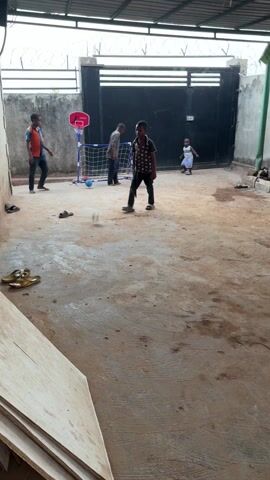 Four young boys are playing outdoors in a fenced-in courtyard. One boy dribbles a soccer ball near the center of the space.  Another boy stands near a small soccer goal with a basketball hoop attached to the top.  A third boy is dribbling a blue ball on the left side of the frame.  A toddler stands near a large gate in the background.  The surface of the courtyard is uneven and sandy-colored.  Sandals and other objects are scattered on the ground. A stack of wooden boards lies in the bottom left corner of the frame. The sky is visible through a partially covered roof above.