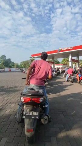 Here is a detailed description of the scene in the video:

The video shows a line of motorcycles waiting to fill up at a Pertamina gas station in what appears to be Indonesia. The person recording is seated on their own motorcycle and is looking towards the front of the queue. The motorcycles are lined up neatly on a paved area. 

Many individuals are visible, mostly on motorcycles. The person filming is wearing a pink shirt, blue jeans, and a black baseball cap. Several others are wearing similar casual attire. The gas station itself is a red and white structure typical of Pertamina stations, with multiple pumps and an overhead canopy.

The sky is blue with scattered white clouds, suggesting a sunny day. There is also some greenery visible in the background, indicating trees and shrubs surrounding the gas station. The scene conveys a routine activity of people getting fuel for their motorcycles at a gas station.