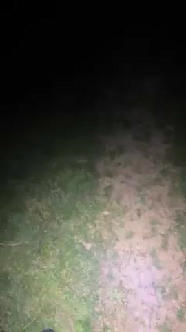 At night, a person is shown walking across a grassy field towards the camera. The person is wearing dark clothing and black boots. They appear to be holding some sort of device. In the distance, lights can be seen.