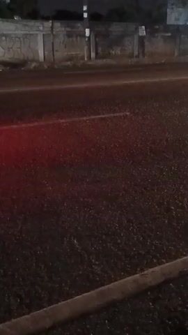 The video shows a nighttime street scene. The camera is positioned low, looking at the road surface. The road appears to be asphalt with a white line marking the lane. There are some small, reflective specks on the road surface, possibly from rain or debris. In the background, there is a concrete wall with graffiti on it, and a pole with a light fixture. A vehicle, appearing red, drives by briefly on the left side of the frame. The lighting is dim, suggesting it's nighttime or heavily overcast.