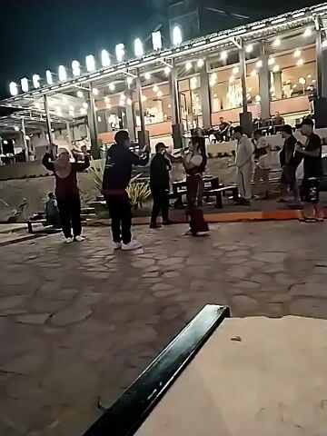 The video shows an outdoor gathering at night. Several people are visible in the foreground and middle ground, some appearing to be interacting or dancing. A building with lights is in the background, suggesting it might be a restaurant or venue. The ground is paved with stones, and there are stairs leading up to a higher level. The lighting suggests it is dark outside, with artificial lights illuminating the scene.