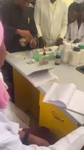 Several individuals are gathered around a laboratory table in what appears to be a learning environment. The main activity involves a person wearing a dark top pouring liquid from a flask into smaller containers arranged on the table. A lit alcohol burner is visible near the containers, suggesting a heating or sterilization process may be underway. Other people around the table, some in white lab coats, are observing the activity, with one person taking notes in a notebook. Several bottles and containers are scattered on the tabletop, adding to the impression that experiments or scientific procedures are being conducted. The setting appears to be a classroom or teaching laboratory.