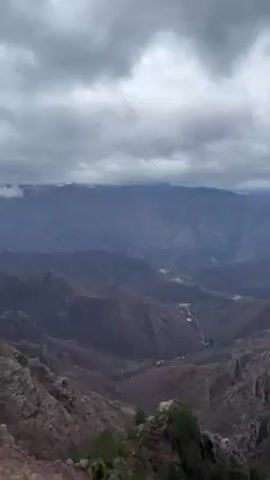 The video shows a view of mountains and valleys under an overcast sky. The mountains are a mixture of brown and gray colors, with some sparse vegetation. In the distance, some mountain peaks have a dusting of snow. The valleys contain a few small clearings and what appears to be a road. The sky is filled with gray clouds, and there is no visible sunlight.