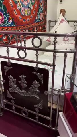 The video shows a traditional bedroom setting. A vintage metal bed frame with ornate details is visible in the foreground. The bed is made with white linens and several white pillows, one of which is embroidered with pink floral patterns. A decorative rug with a vibrant floral design in shades of red, pink, green, and blue hangs on the wall above the bed. To the right of the bed, on a shelf, is a small bust of a person. The overall impression is of a preserved historical room.