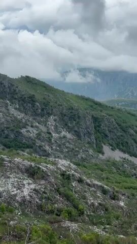The video shows a mountainous landscape with green vegetation and rocky terrain under a cloudy sky. The mountains are covered with trees and shrubs, with areas of exposed rock. Clouds obscure the peaks of the distant mountains. The camera angle is looking down on the landscape.