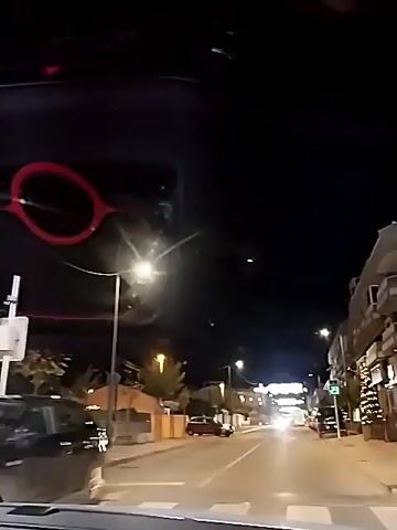 The video shows a night-time scene from inside a moving car driving on a city street. Streetlights illuminate the road and buildings on either side. There's a Christmas tree decorated with lights in front of a building on the right. Several cars are parked along the street. An alarm is blaring loudly in the background.