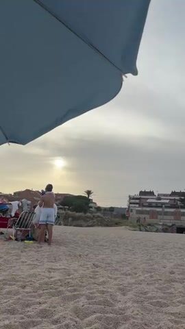 The video begins with a close-up of a blue beach umbrella, partially obscuring the view. The camera pans slightly to the right, revealing a sandy beach with a few people scattered across it. In the background, there are buildings and vegetation. The sun is visible in the sky, peeking through the clouds. The scene appears to be a beach in the late afternoon or early evening, as the lighting is soft and diffused. A young man in white shorts stands on the beach, facing away from the camera. A group of people are gathered further down the beach, some sitting on chairs and blankets. The camera continues to pan, showing more of the beach and the buildings in the distance. There are a few more people on the beach, some sitting together and others walking. The video ends with a wide shot of the beach, with the buildings and the sky filling the background.