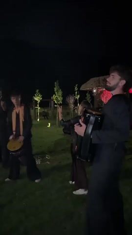 A group of musicians perform outdoors at night. In the foreground, a man with a beard plays an accordion. To his left, a woman sings into a microphone. Further left, a young man in a dark outfit with an orange and yellow sash plays a frame drum. To the far left, another person plays a flute. The musicians are on a grassy area, with trees and a building in the background. Red lights are visible on the building. The scene is lit by artificial lights and the natural darkness of night.