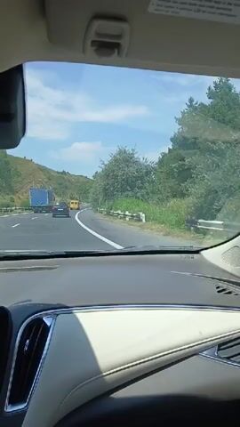 The video shows a view from inside a car driving on a highway. The dashboard of the car is visible in the foreground. Outside the car, there are other vehicles on the road, including a truck and a smaller vehicle. The road is surrounded by trees and greenery, and the sky is blue with some clouds. The sun is shining. The video appears to be taken during the daytime.