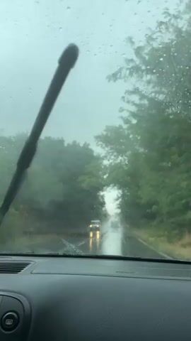 The video shows a view from inside a car driving on a wet road during a rainstorm. Raindrops are visible on the windshield, obscuring the view of the road ahead, which is lined with trees on both sides. Another car can be seen in the distance, its headlights reflecting on the wet pavement. The car's windshield wipers are intermittently activated to clear the view. The interior of the car, including the dashboard and air vents, is partially visible.