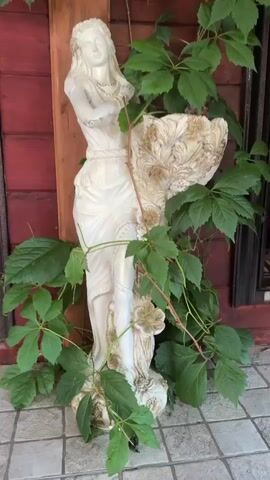 A statue of a woman adorned with a necklace stands amidst lush green vines that twine around her and a planter she holds. The statue and vines are positioned in front of a dark red wooden wall. The ground beneath is covered with light-colored square tiles. The video begins with a close-up of the statue's lower body and the vines, then pans upwards, revealing more of the statue and the wall, finally ending with a view of the vines reaching towards electrical conduits and wiring attached to the wall.