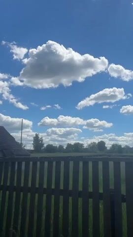 The video captures a view of a bright blue sky filled with fluffy white clouds. In the foreground, a dark wooden fence is visible, with hints of green grass beyond it. In the distance, a line of trees forms a backdrop, and a roofline of a building can be seen to the left. The overall impression is a sunny day in a rural or suburban setting.