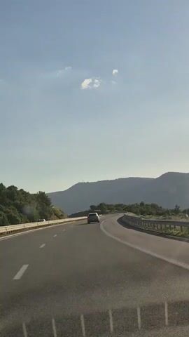 The video begins with a view of a highway as seen from a moving vehicle. The road curves to the right, with guardrails on either side. In the distance, mountains are visible under a clear blue sky with a few wispy clouds. A single white car is seen ahead on the highway. As the video progresses, the sun becomes more prominent, shining brightly and creating lens flare effects on the camera. The mountains in the background are still visible, but become more obscured by the intense sunlight. The final frames show a close-up of the sun and the sky, with a blurry foreground that appears to be the inside of the vehicle.