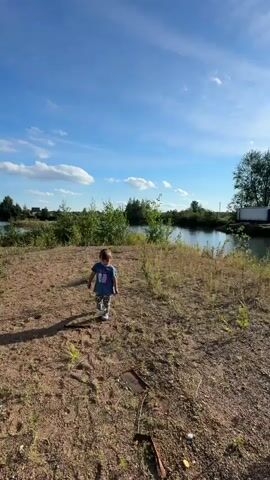 A young child with brown hair, wearing a blue t-shirt and grey pants, walks and then runs across a dirt and patchy grass area. In the background, there is a body of water, trees, and a bright blue sky with scattered white clouds. The sun is casting shadows from the trees and the child.