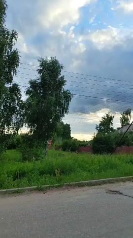 This media file depicts a scene in Tsibino, Russia. The viewpoint is from a roadside, looking across a grassy verge towards a residential area. Several tall, leafy trees, likely birch, dominate the foreground and midground, with power lines strung between them. Beyond the trees, a red-colored house with white trim and multiple windows is visible, as is a wooden fence. Further down the road, more houses with red roofs can be seen, along with scattered trees and more greenery. The sky is a mix of blue patches and heavy, grey clouds, suggesting a dynamic weather pattern, possibly approaching rain or a storm. The lighting indicates it is likely late afternoon or early evening, with the sun attempting to break through the clouds. The road itself appears to be asphalt with some wear and tear, including potholes. There is no visible text in the media file. The overall impression is of a semi-rural or suburban setting in Russia during overcast weather. No people or specific activities are clearly depicted, suggesting a general view of the landscape and residential area.