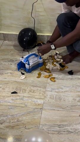 A man in a white t-shirt, jeans, and a blue baseball cap is squatting on a marble-patterned tile floor. He is barefoot and using a blue and white electric air pump to inflate small gold and white balloons, which are scattered around him. A single black balloon sits nearby. The scene takes place indoors, against a cream-colored wall with a power cord running along it. The man is focused on inflating the balloons, his hands manipulating them onto the pump nozzle.
