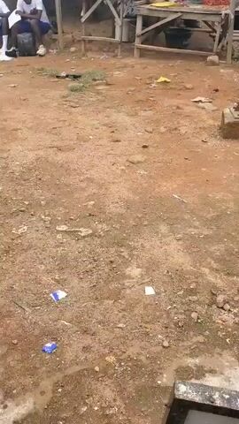 The video shows an outdoor scene with a dirt ground. There are several pieces of trash scattered on the ground, including blue and white wrappers. In the background, there are some wooden structures that appear to be market stalls. There are also a few people visible in the background. One person is sitting down wearing a white shirt and blue shorts, with a black backpack next to them. Another person is standing further back. The overall impression is of a somewhat unkempt outdoor area, possibly a market or a public space.