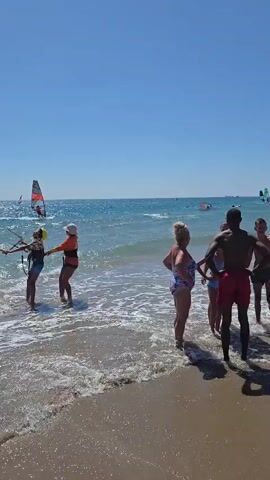 A sunny day at the beach with people in and near the water. On the left, two individuals, one wearing an orange life jacket and a white hat, are standing in the shallow water, holding onto a windsurfing sail attached to a board. Another person is visible further out in the water with a windsurfing sail. In the center and right of the frame, a group of people are standing on the sandy shore or wading in the water. One woman in a patterned swimsuit is facing away from the camera. A man with dark skin is in the foreground, wearing red shorts, and appears to be looking towards the water. Other individuals are standing behind him. The sea is visible with waves and several more windsurfing sails can be seen further out on the horizon. The sky is clear and blue.