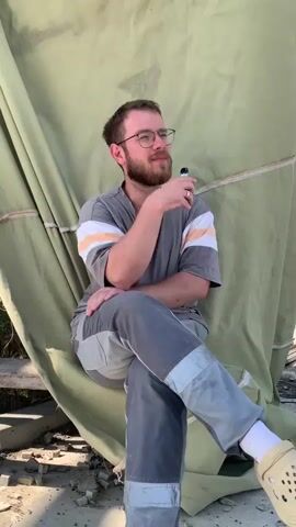 A man with a beard and glasses is sitting in front of a large, draped piece of olive-green fabric. He is wearing a grey t-shirt with white and orange stripes on the sleeves, and grey work pants with lighter grey patches. His legs are crossed, and he is holding a dark object in his right hand. He is looking upwards with a thoughtful expression. The fabric behind him is creased and appears to be part of a larger structure or shelter. The ground beneath him is littered with debris, possibly from a construction site or a damaged area. In the background, a hint of blue sky and a distant body of water can be seen, suggesting an outdoor setting under daylight. The lighting suggests it's daytime, likely morning or afternoon. The overall impression is one of contemplation or waiting in a somewhat utilitarian or desolate environment. The man appears calm and reflective. There is no visible text in the media.
