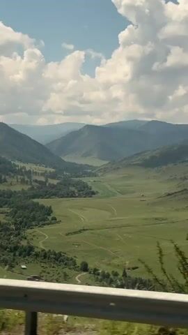 The video shows a view from a moving vehicle looking out at a mountainous landscape. In the foreground, there is a silver metal guardrail. To the left, a large green tree with sparse leaves is visible. The landscape beyond consists of rolling green hills and mountains under a cloudy blue sky. Further into the distance, the mountains appear steeper and covered in trees. A signpost with a red and white curved arrow indicating a sharp turn is also visible on the right side of the frame. The camera pans slightly, capturing the changing scenery of the road and the natural environment.
