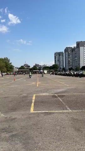 The video shows an outdoor motorcycle training or testing area. The ground is paved with asphalt and marked with yellow lines, indicating lanes and practice zones. Several orange traffic cones are placed on the ground, delineating the course. In the background, there are several buildings, including apartment complexes and what appears to be a mosque with a minaret. There are also trees and some temporary structures with tents. Multiple people are present, some standing and observing, while others are on motorcycles. At least two individuals are actively riding motorcycles within the marked area, navigating through the cones. A line of parked motorcycles is visible in the middle ground, suggesting this is a designated area for motorcycle activities. The sky is blue with scattered clouds, indicating fair weather. The overall scene depicts a structured environment for motorcycle practice or assessment.