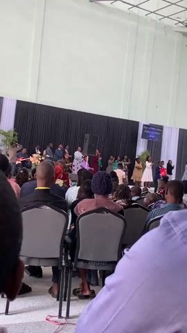 The video shows a crowd of people seated in a large room, facing a stage. On the stage, a group of people are gathered, some are standing and singing, while others are seated behind them. There are musical instruments visible, including a guitar, and a speaker is placed on the stage. A banner is hung on the back wall of the stage with text that is difficult to read. The audience members are mostly facing the stage, with some looking at their phones. The room appears to be a place of worship or a gathering.